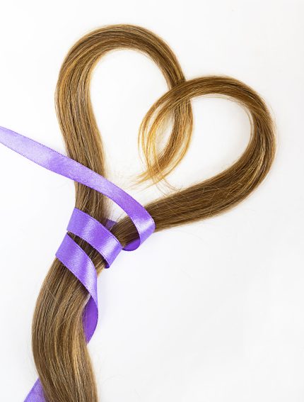 long blond hair donation put like heart with lavender ribbon as symbol of fight against cancer on white background. Top view. Natural material for making wig for patients. Vertical, isolated.